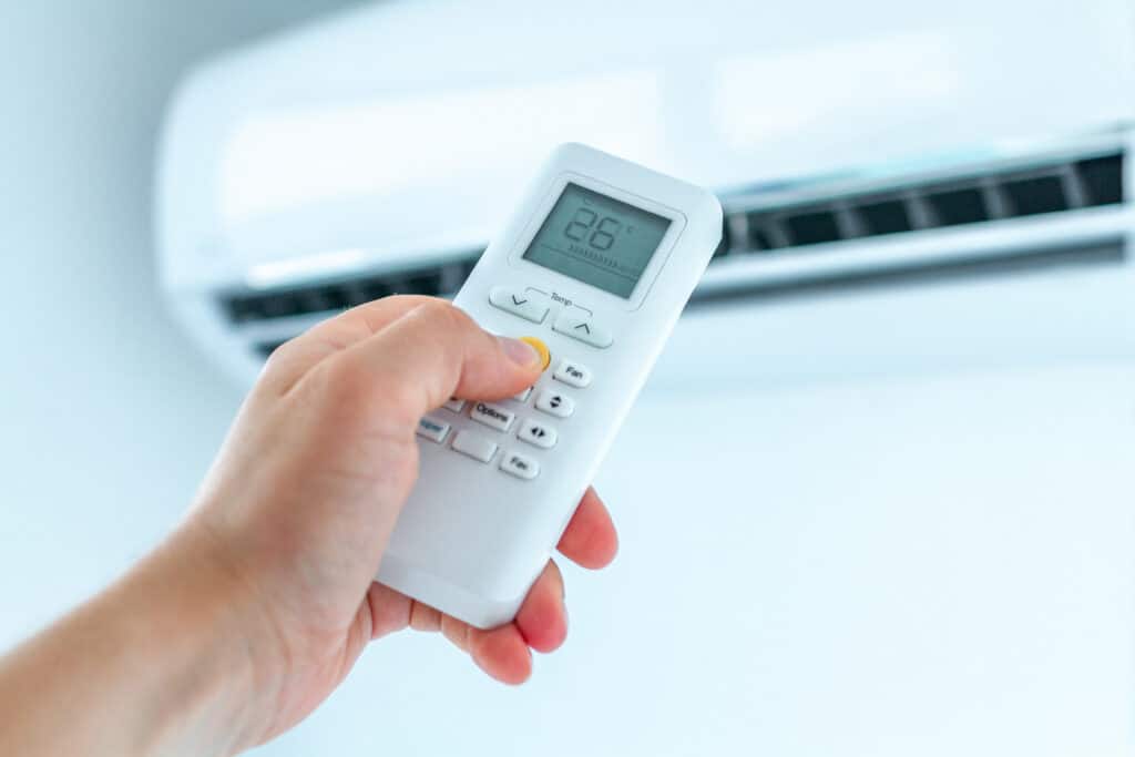 Main presse le bouton jaune d'une télécommande de climatiseur, affichant 26°C en mode AUTO. Unité AC en arrière-plan.