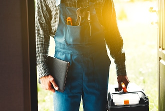 Technicien en salopette bleue et chemise, outils dans les poches, tenant une boîte à outils et un dossier, entrant dans une maison.