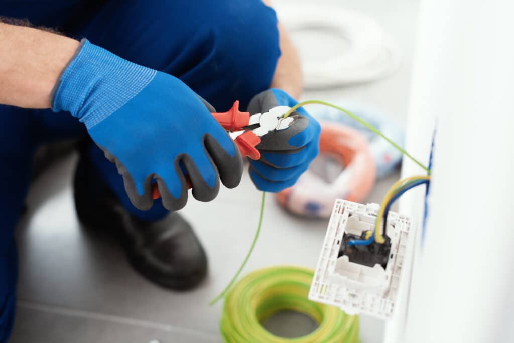 Électricien en gants bleus coupant un fil électrique vert et jaune avec des pinces, installant un câblage mural près d'une prise.