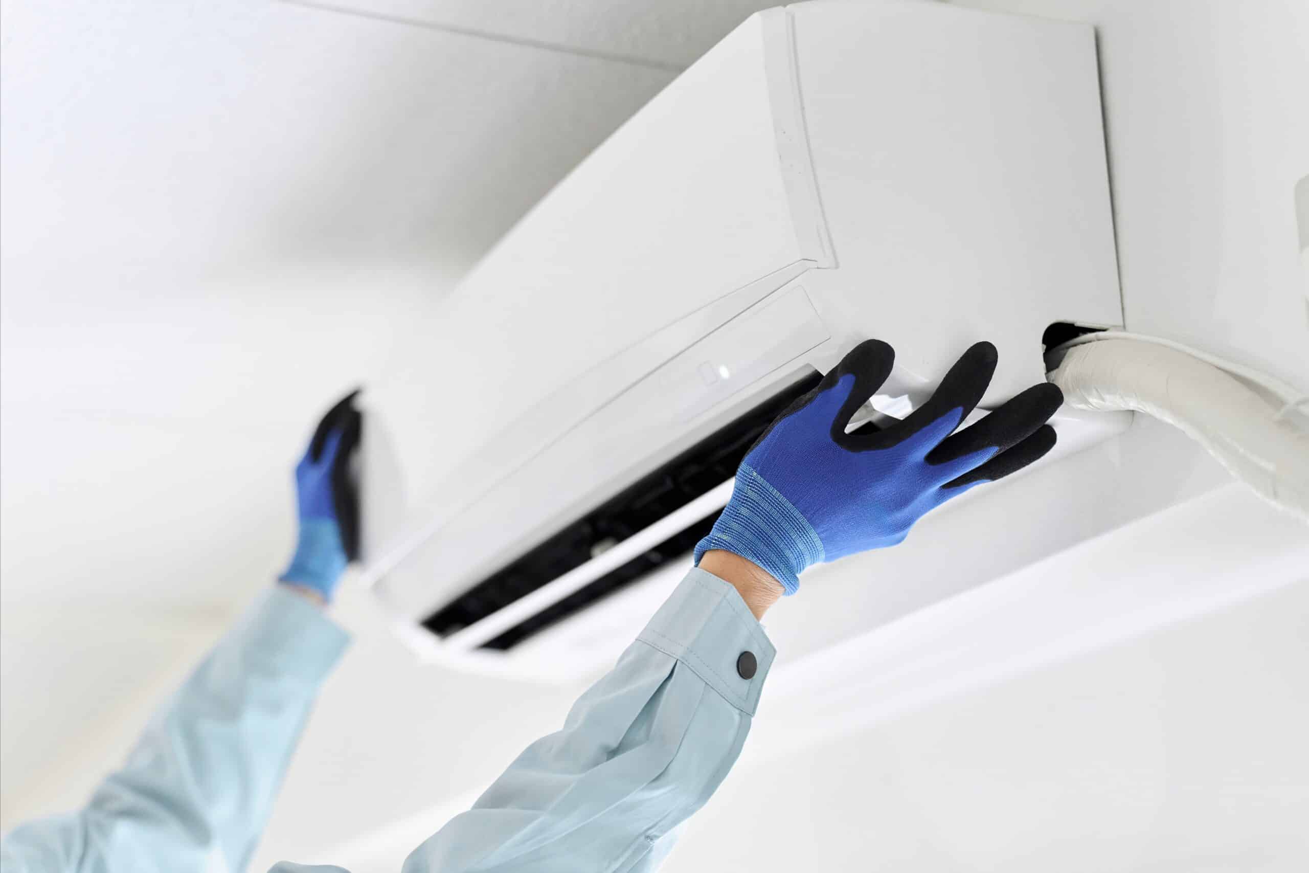 Technicien en gants bleus et noirs installant ou entretenant un climatiseur mural blanc. L'appareil est ouvert, révélant conduits et isolation.