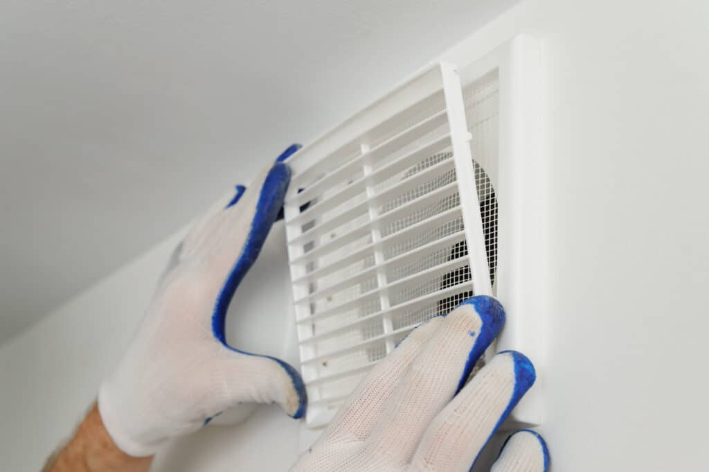Un ouvrier, mains gantées, fixe une grille de ventilation blanche à persiennes et moustiquaire sur un mur clair. Installation ou entretien CVC.