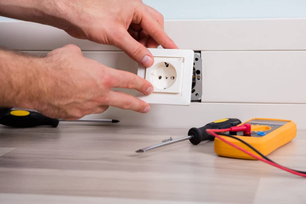 Mains d'électricien installant une prise électrique murale blanche. Des tournevis et un multimètre jaune sont posés sur le sol en bois.