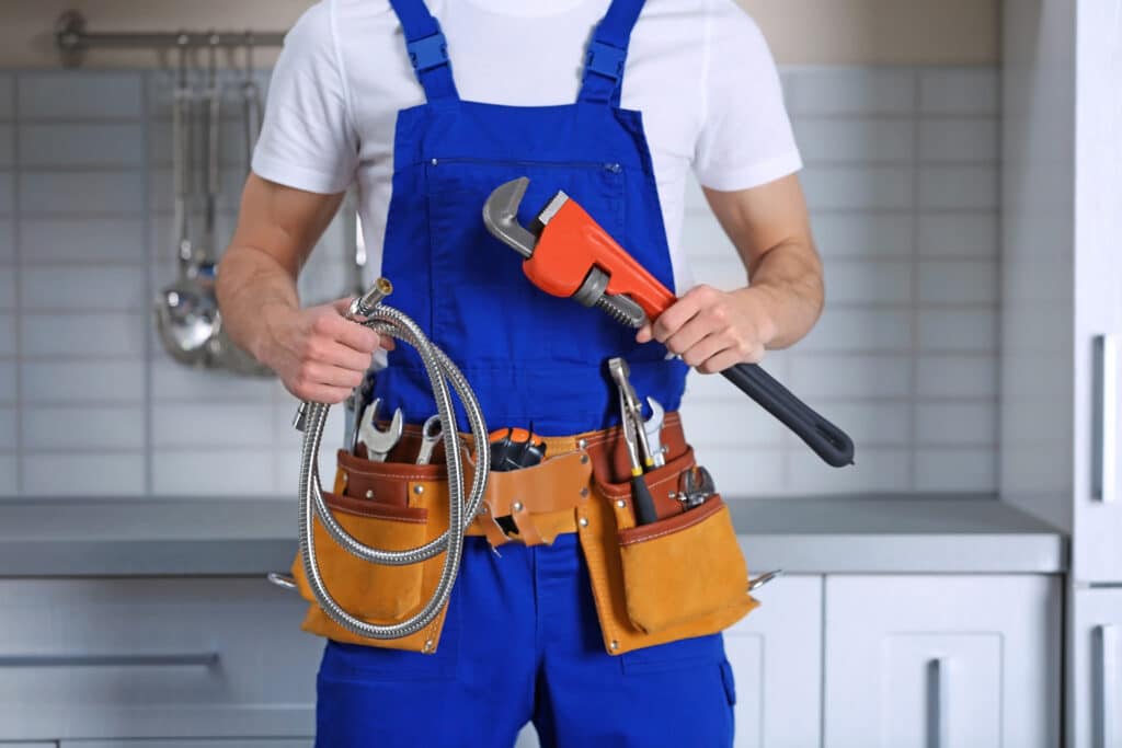 Plombier en salopette bleue et t-shirt blanc, tenant une clé serre-tube orange et un flexible. Ceinture à outils garnie en cuisine.