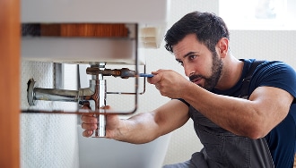 Plombier barbu concentré répare les tuyaux d'un lavabo de salle de bain avec une clé, tenue d'artisan.