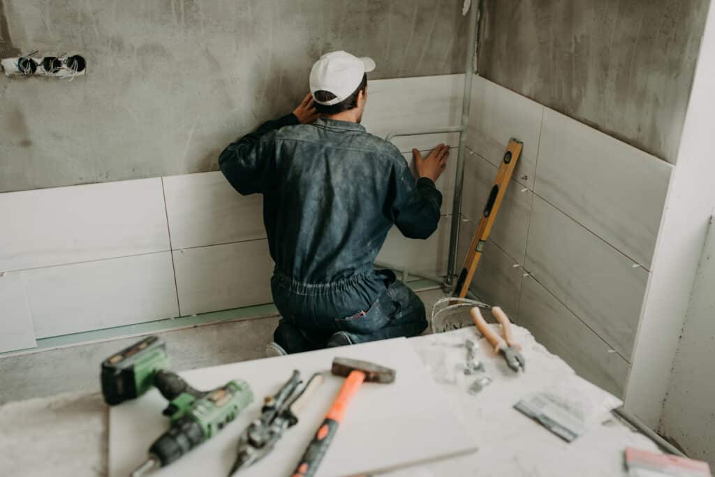 Ouvrier en salopette bleue et casquette blanche installant des carreaux de faïence blancs sur un mur brut. Des outils de carrelage sont au premier plan.