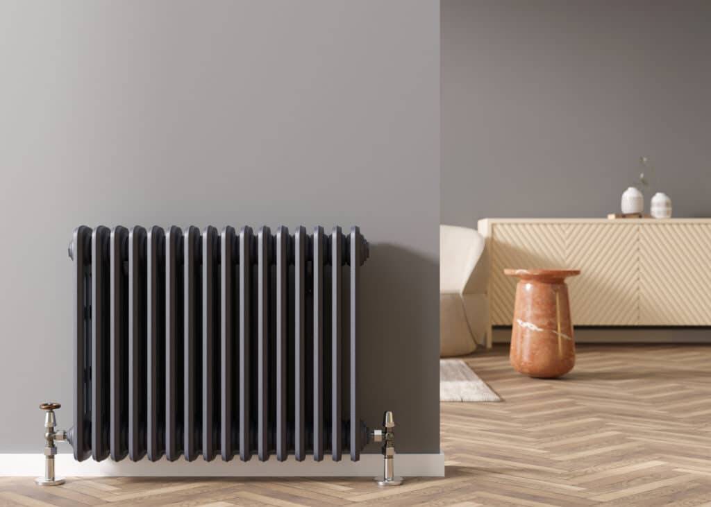 Radiateur en fonte gris anthracite avec robinets vintage sur parquet chevrons. Salon moderne, meuble bas texturé, vase marbre.