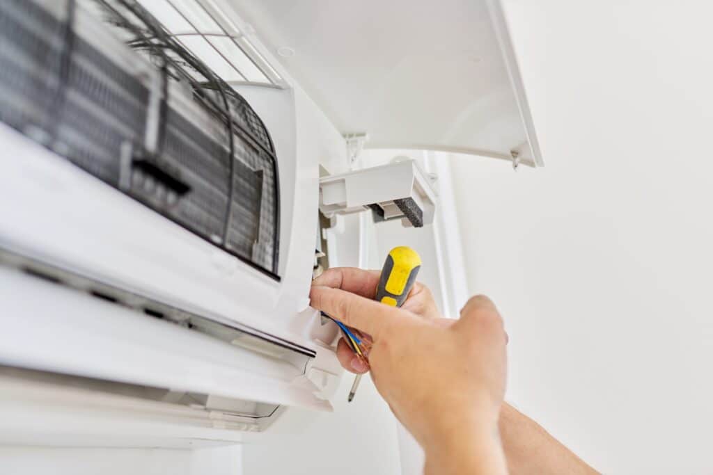 Technicien réparant un climatiseur blanc mural, mains connectant des fils électriques colorés avec un tournevis, composants internes visibles.