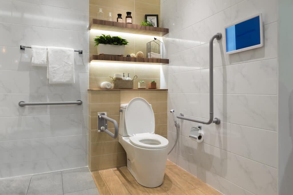 Salle de bain moderne et accessible PMR avec toilettes, barres d'appui, étagères éclairées, carrelage blanc marbré et décor chic.