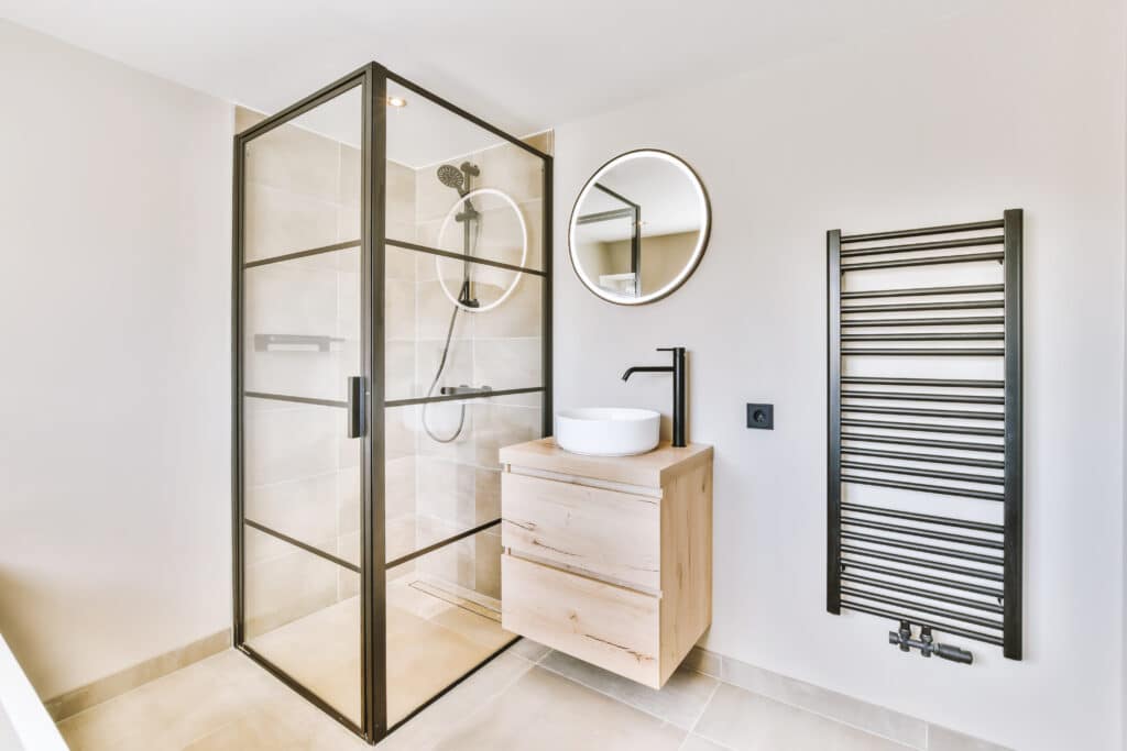 Salle de bain moderne et épurée: douche à l'italienne avec cadre noir, meuble lavabo en bois clair, miroir rond éclairé et sèche-serviettes mural noir.