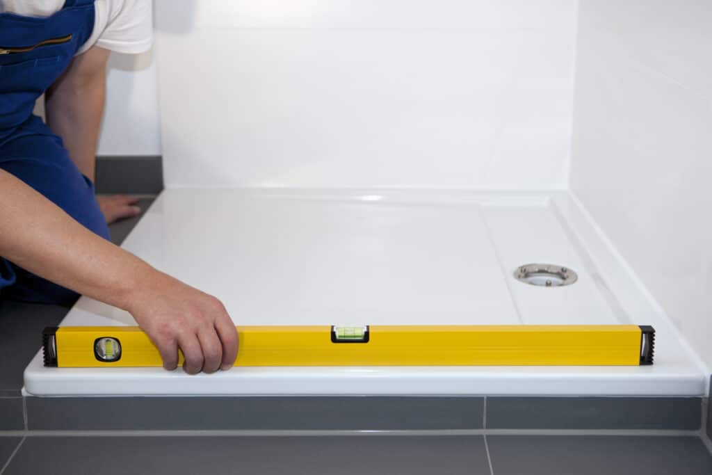 Plombier vérifiant le niveau d'un receveur de douche blanc avec un niveau à bulle jaune, installé sur un carrelage gris. Travaux de salle de bain.